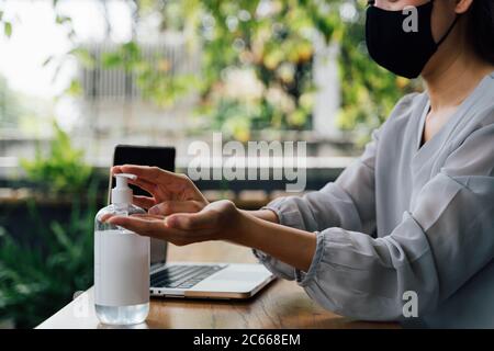 Side view of young Asian female in protective mask applying sanitizer on hands while sitting at table on terrace during pandemic Stock Photo