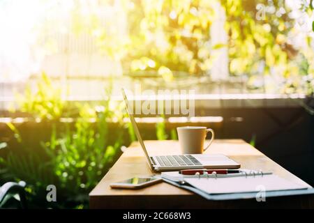 Modern laptop and smartphone placed on table near open planner with pen and cup of hot beverage on sunny summer day on terrace Stock Photo