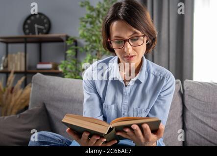 Interested woman in eyeglasses reading book at home Stock Photo