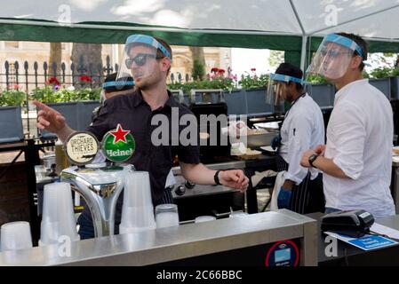 With the Coronavirus pandemic lockdown rules being eased, pubs have now re-opened and staff working under cover outside the riverside Trafalgar Tavern, a pub on the Thames at Greenwich, serve customers wearing required face shields, on 5th July 2020, in London, England. Stock Photo