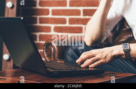 a man is holds his hand on the laptop's touchpad. He sits on a brown armchair in his own cabinet Stock Photo