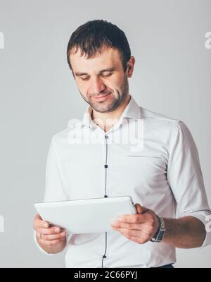 a businessman smiles and holds a white tablet in his hands. A man is standing in luxurious shirt on a white background. Stock Photo