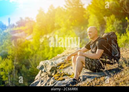 a traveler with a backpack on his shoulders sits on a rock in the background of the forest in the summer. A tourist looks at the nature around the roc Stock Photo
