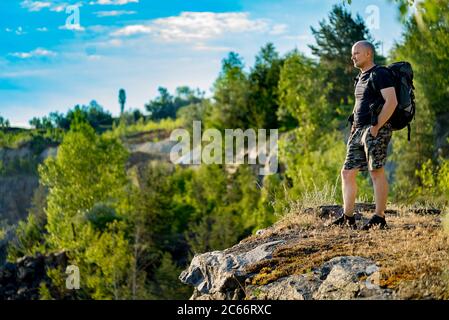 tourist with a backpack on his shoulders stands on a rock in the background of nature in the summer. A traveler looks at the landscape in front of him Stock Photo
