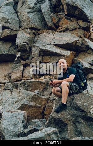 a traveler with a backpack behind his shoulders sits on the rock at the foot of a high cliff. A traveler holds a small pebble in his hand for the memo Stock Photo