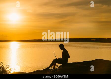 a man works behind a laptop on the background of sunset. The guy combines rest with work Stock Photo