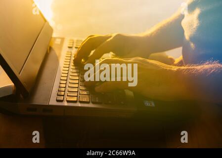 a man types the text on the keyboard of his laptop at sunset near the river. The guy busy with work Stock Photo