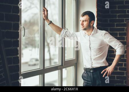 businessman looks through a window from his office. A man in business clothes stands in the room and leans his hand on a window frame Stock Photo