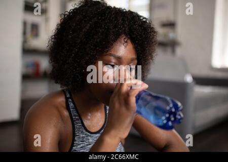 Tired black female sipping water from bottle during break in training at home