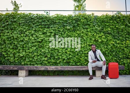 businessman sitting on a bench with a red suitcase and mobile phone in hand. business man traveling. Stock Photo