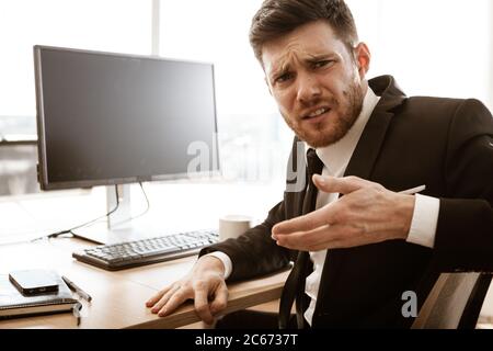 Business crisis concept. Emotional young businessman at work. Manager sitting at the office table shows worried face and jiggles his hand. Busy man in Stock Photo