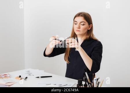 Portrait of young beautiful lady sitting at the white desk and taking photos of cool notes on her cellphone isolated Stock Photo