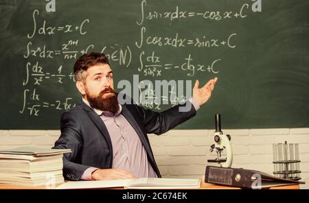 Talking to students or pupils. School teacher concept. Teacher bearded man tell interesting story. Teacher charismatic hipster sit at table classroom chalkboard background. Educational conversation. Stock Photo