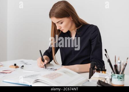 Portrait of young nice lady sitting at the white desk with open book while writing notes on paper isolated Stock Photo