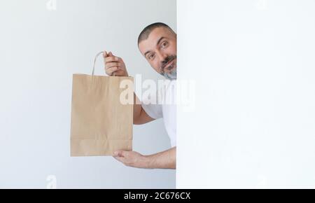 Handsome smiling man with beard peeking behind a white wall and holding paper bag in hands isolated on white background Stock Photo