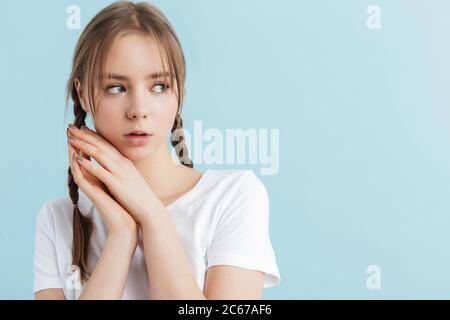 Portrait of young beautiful girl with two braids in white t-shirt holding hands near face while dreamily looking aside over blue background Stock Photo