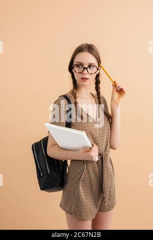 Young beautiful lady with two braids in tweed jumpsuit and eyeglasses with black backpack on shoulder holding book and pencil in hand near head Stock Photo