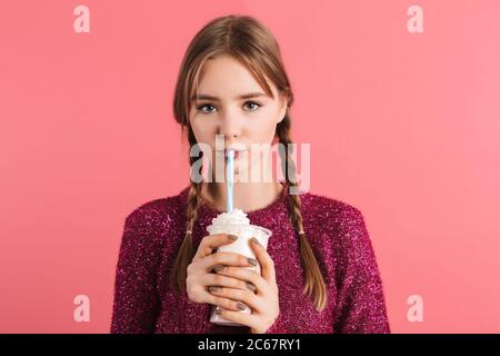 Young pretty girl with two braids in sweater drinking milkshake thoughtfully looking in camera while spending time over pink background Stock Photo