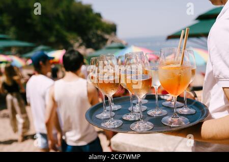 A waiter with a tray in hand carries cocktails and drinks at a party on the beach - an orange cocktail in a large glass with a tube Aperol spritz and Stock Photo