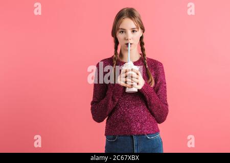 Young dreamy girl with two braids in sweater and jeans drinking milkshake thoughtfully looking in camera over pink background isolated Stock Photo