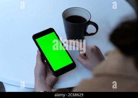 over should of woman tapping green screen smartphone at cafe. A cup of coffee on white table. Blur background Stock Photo
