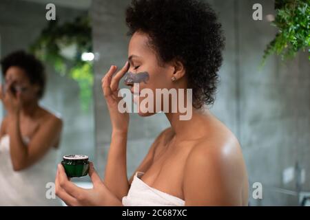 Side view of happy ethnic lady smiling and smearing mud on face in bathroom at home Stock Photo