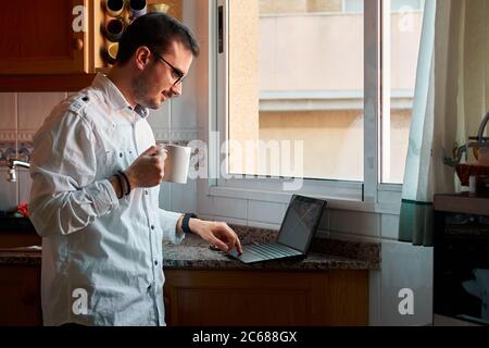 Young man looks at his laptop while drinking coffee in his kitchen Stock Photo
