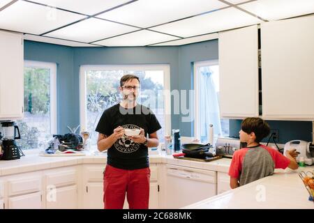 Father stands in the kitchen eating a bowl of cereal near son Stock Photo