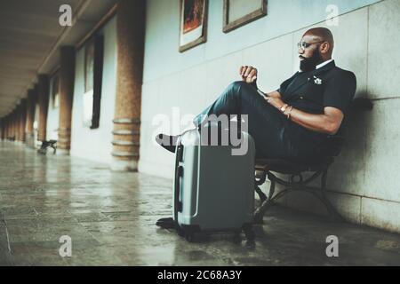 A mature African bald bearded businessman is waiting for a train to start his business trip, looks at the clock while sitting on a wooden bench at the Stock Photo