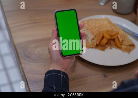 over shoulder of hand holding green screen smartphone. Blur food in plate on wooden table. Stock Photo