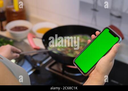 over shoulder of woman using green screen smartphone while cooking in kitchen. blur background Stock Photo
