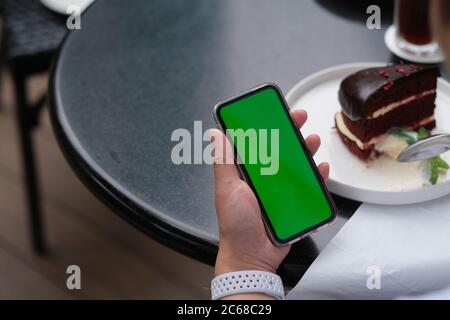 over shoulder of people holding green screen smartphone. blurred chocolate cake on black table. Stock Photo