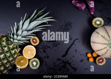Pineapple, honeydew melon, green and red kiwis, Barbary figs, oranges and lemons on a wet black background. Stock Photo