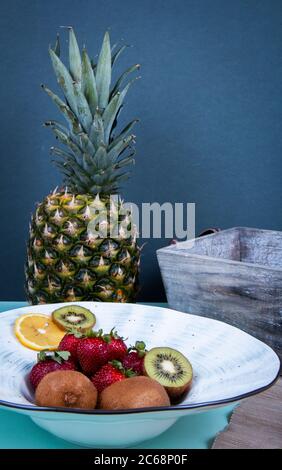 Kiwifruits, strawberries, lemon and orange section in ceramic dish and a pineapple in front of a gray wall. Stock Photo