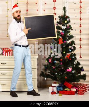 Guy near Christmas tree on wooden wall background. Celebration and New Year mood concept. Man with beard holds blank blackboard and point at it, copy space. Santa Claus in red hat with shocked face Stock Photo