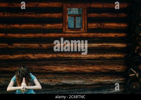 Girl with bowed head and hands in namaste on the background of an old brown log wall. The concept of loneliness, old age, humility. Stock Photo