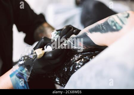 Close up professional tattooer in black gloves doing tattoo on hand using tattoo machine in studio Stock Photo