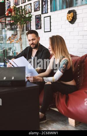 Professional tattoo master sitting on sofa with girl discussing sketch of new tattoo in tattoo studio Stock Photo