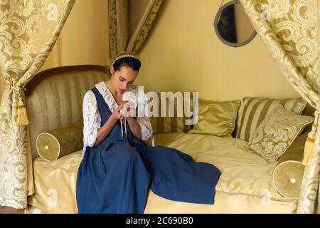 Dutch Master style portrait of a beautiful young woman in authentic renaissance costume lying on a luxury antique canopy bed in a golden bedroom of a Stock Photo