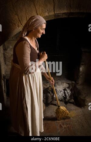 Woman dressed in renaissance maid costume using her broom to clean the fireplace in the kitchen of a property released authentic medieval castle in Fr Stock Photo