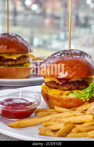 Two hamburgers with beef cutlet, cheese, french fries, tomato, salad, sauce and ketchup on a white plate and a light wooden background Stock Photo