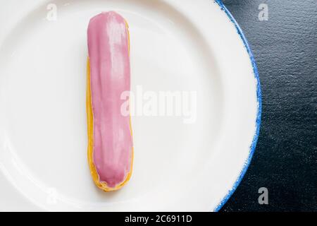 Eclair with pink glaze on a white plate. Raspberry eclair close up in the glaze on a black wooden table. Top view. Stock Photo