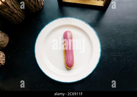 Eclair with pink glaze on a white plate. Raspberry eclair in the glaze on a black wooden table. Top view. Stock Photo