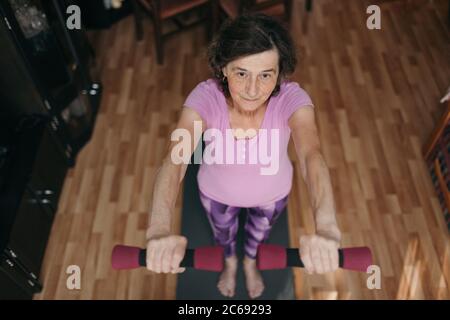 Retired woman exercising with dumbbells at home. High angle shot of 70 years old woman in purple sportswear exercising shoulders holding dumbbells bot Stock Photo