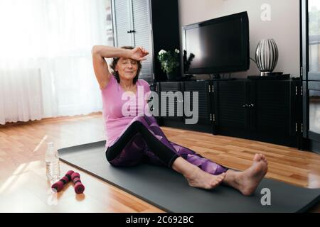 Tired elderly woman after exercise at home. Weary senior woman in purple sportswear sitting on fitness mat wipes sweat from her forehead after workout Stock Photo