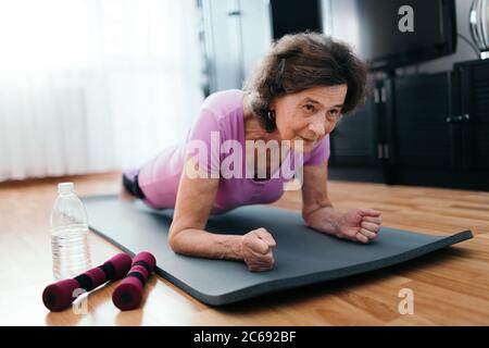 Senior woman workout at home. Low angle shot of elderly woman at her 70s lying on fitness mat exercising plank indoors.