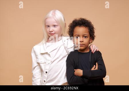 two friendly black afro american and albino children stand together isolated over beige background, diverse positive boy and girl stand hugging. child Stock Photo