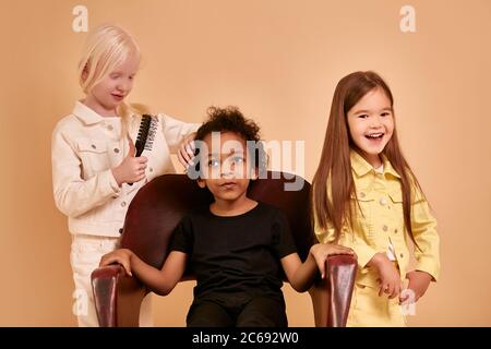 albino child girl combing curly hair of african boy isolated, girl brushing hair of cute serious boy sitting on chair. caucasian girl with long hair s Stock Photo