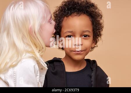 friendly diverse children play, tell stories and secrets to each other isolated in studio. girl with albinism syndrome share secret with african boy Stock Photo