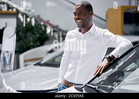 young africanamerican man came to see automobiles in dealership or cars showroom, he stands next to business class auto Stock Photo
